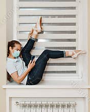 A female ballerina in a mask, in everyday clothes, in jeans and a t-shirt, in Pointe shoes at home on the windowsill doing