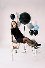 Happy emotional glamour smiling young woman in black dress and shoes with birthday holiday ballons sitting on white