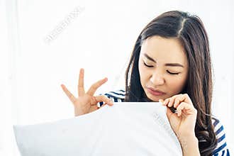 Beautiful woman sniffing a white pillow with a foul odor or musty smell, the face showing disgusting symptoms at white bedroom,