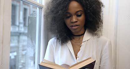 The young attractive afro-american woman is reading the book on the window-sill. Close-up portrait.