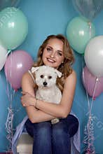 Charming, pretty, cheerful girl with beaming smile, curly hair holding dog on arms, celebrating pet`s birthday, animal