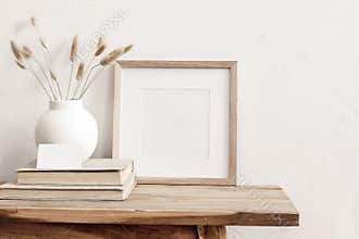 Square wooden frame mockup on vintage bench, table. Modern white ceramic vase with dry Lagurus ovatus grass, books and
