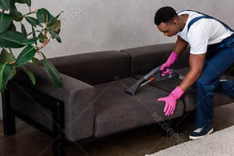 Focus of african american cleaner in uniform cleaning upholstery of couch at home