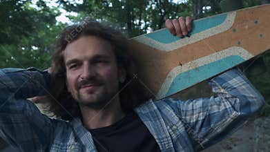 Portrait: A young hipster guy throws a longboard around his neck. He looks into the camera. It is located in the forest