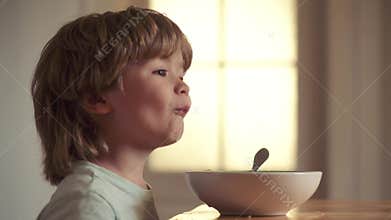 Portrait of sweet little laughing baby boy with blonde hair eating from plate holding spoon close-up. Little boy sitting
