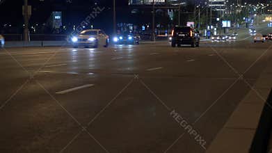 Autos with lit headlights drive along street in night city