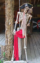 Pirate skeleton with peg leg and eye patch and hat and dead parrot on shoulder propped on tree trunk pillar with pirate flag in
