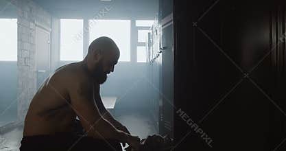 Young athletic shirtless Caucasian man sitting alone in dark gym locker room resting after workout, overcoming trouble.
