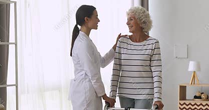Woman caregiver helping happy old disabled grandmother stand hold walker