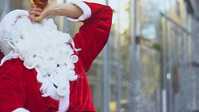 Bad drunk Santa Claus sits near a city office building. Santa Claus drinks alcohol from a bottle on the street