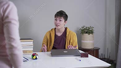 Back view of adult Caucasian woman closing son`s laptop and pointing at stack of books lying at the table. Addicted