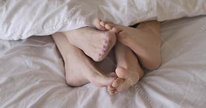 Close-up top view of senior female Caucasian feet touching male legs in bed, both pairs hiding under white blanket