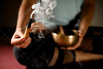 Girl in sport clothes practicing yoga in a studio room sitting in lotus pose torching the palo santo