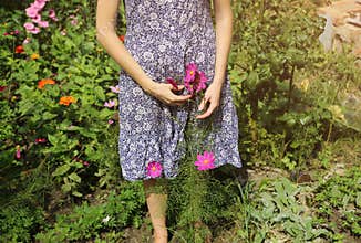 Beautiful girl in old rustic clothes is holding pink flowers near the house. Summer day on farm barn yard. Natural vintage woman