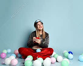 Teenager in reindeer pajamas, red socks, sleep mask. Holding cup, dreaming, sitting on floor, balloons, blue background. Close up