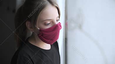 Sad girl wearing a cloth face mask looking through window during worldwide virus epidemic.