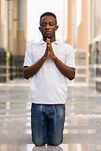 Portrait of young African man kneeling and praying with eyes closed outdoors