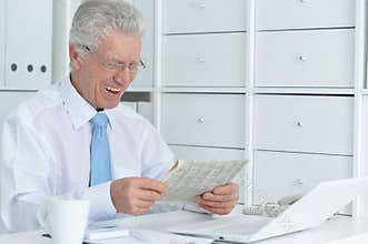 Beautiful old caucasian man with laptop reading newspaper