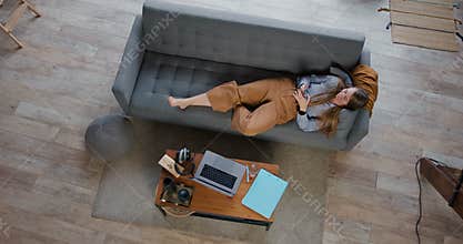 Top view young beautiful relaxed blonde woman lying down on cozy sofa using smart phone social network app at home.