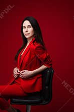 Portrait of caucasian female with long dark straight hair in red office suit, black shoes sits on black chair