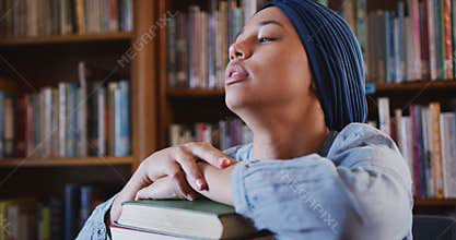 Asian female student wearing a blue hijab sitting and leaning on a pile of books and thinking