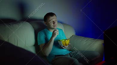 A lonely man spends a boring evening watching TV sitting on a sofa and eating chips
