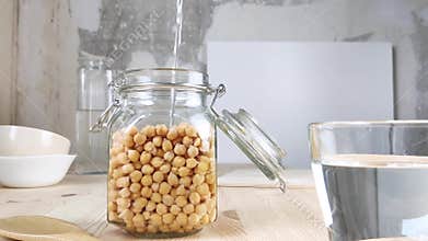 Chickpeas in transparent glass jar are filled with water. Soak beans for further cooking. Healthy vegan food. Side view, wooden
