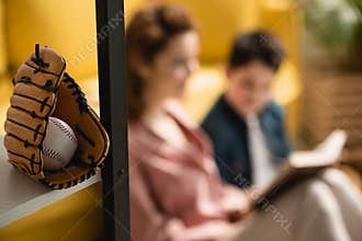 Focus of baseball glove and ball near mother and son sitting on floor and reading book together