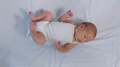 Newborn baby girl in bed. Top view of lovely infant in white pajama with open mouth lying on bed moving arms and legs