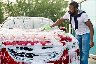 Young attractive African American man washing his red luxury car in a self-service car wash station outdoors with