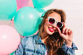 Adorable brunette young woman looking through stylish sunglasses and posing with smile next to colorful balloons. Close