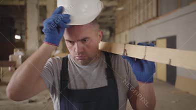 Portrait of a tired male caucasian worker walking with wooden plank at the construction site. Adult builder taking off