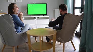 Man and woman are sitting in chairs, watching TV with a green screen, discuss what they saw and switching channels with