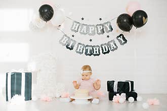 A one year old baby in a pink dress tries the first cake in her life on a black and white photo zone with a happy birthday