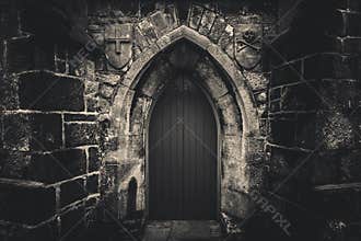 Scary pointy wooden door in an old and wet stone wall building with cross, skull and bones at both sides in black and white.