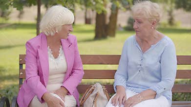Nice mature caucasian women sitting at the bench in the summer park. Senior woman with blond hair complaining the pain