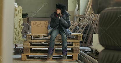 Little Syrian refugee in dirty warm clothes sitting on pallets at the construction site with Help cardboard. Lonely