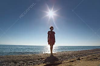 Woman stands ashore opposite sun