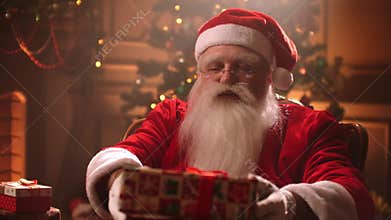 An elderly man with a white beard and a Santa Claus costume Hands a gift to the camera and smiles