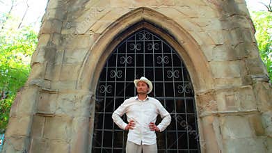 A man in a white straw hat stands in front of an old Gothic building and turns his head thoughtfully. Quest. Crypt