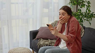 Young caucasian woman sitting on the sofa using smartphone reading good new and making surprised emotion on her face