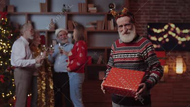 Senior Male Showing New Year Gift while His Friends Celebrating Standing on Decorated Background at Home
