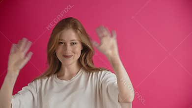 A young european or american ginger woman in a white T-shirt is posing in the studio on a pink background. A happy and
