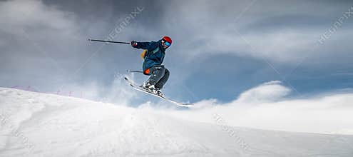 A sportsman skier in ski equipment jumps down a steep snowy slope of a mountain against the backdrop of a blue sky and
