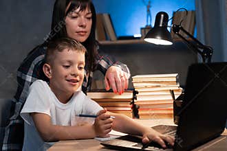 mom helping son with homework while sitting at desk at home using laptop and books at night. e-learning, homeschooling