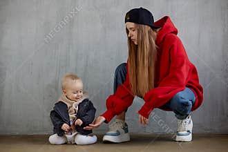 Young long haired mom in red hoodie and black baseball cap plays with her cute baby in beige hoodie and funny motorcycle