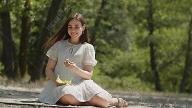 A happy girl with beautiful smile in positive emotions sits on the sand in a summer forest. A young lady in a white
