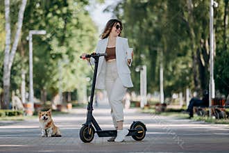 Beautiful young woman in white suit and sunglasses standing with her electric scooter and Corgi Dog in city parkland and drinking