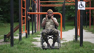 Wide shot portrait of positive brave soldier in wheelchair listening to music in headphones smiling. Courageous African