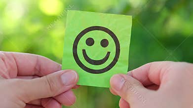 A woman holding a green paper note with a happy face on it in her hands against a background of foliage.